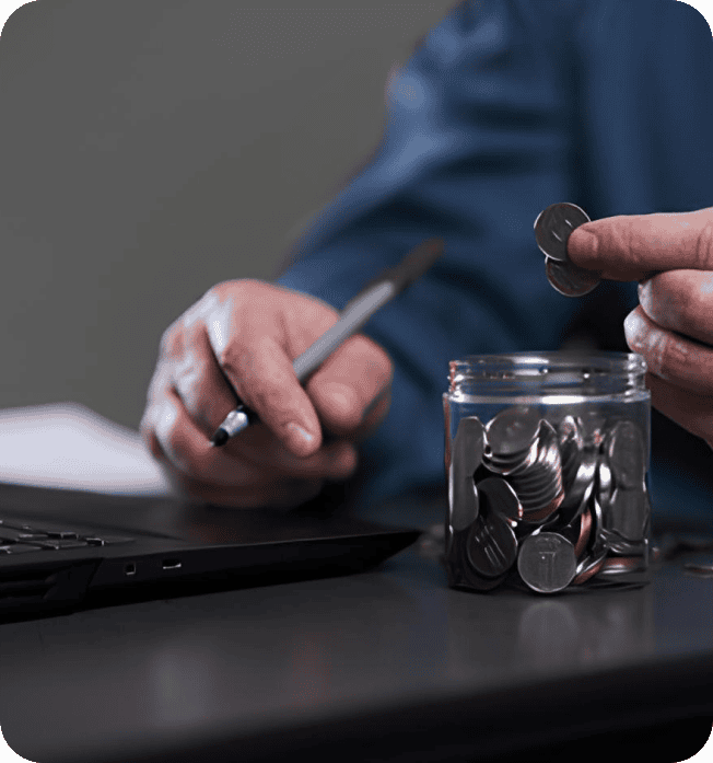 Coins being inserted into savings jar with laptop and pen in background.